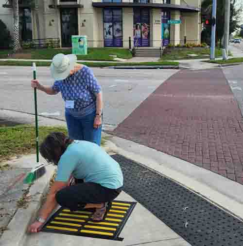 Street view of same intersection, in foreground is a 2’ x 2’ temporary Tactile Direction Indicator (TDI) with a black background and yellow bars perpendicular to the line of travel indicating the correct crossing alignment.  In the background, 2 women install a second TDI, Jen Graham, COMS is squatting with her back to the camera, and Beezy Bentzen, COMS, stands in front of her looking down at the TDI.